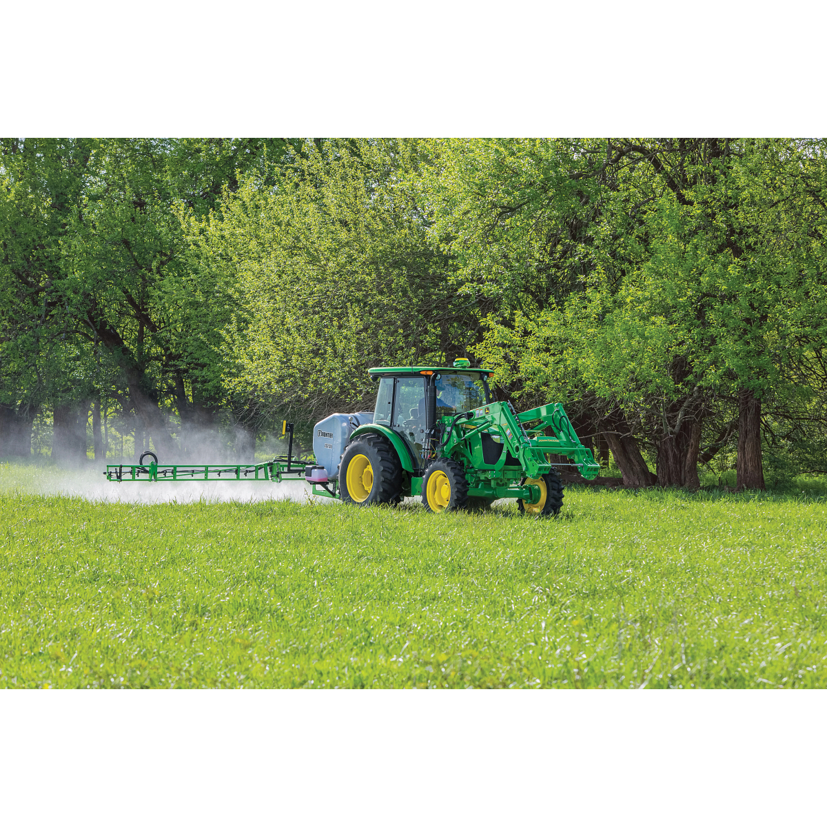 A tractor with a front loader and a wide boom sprayer drives across a grassy field, spraying mist while trees with spring foliage line the background.