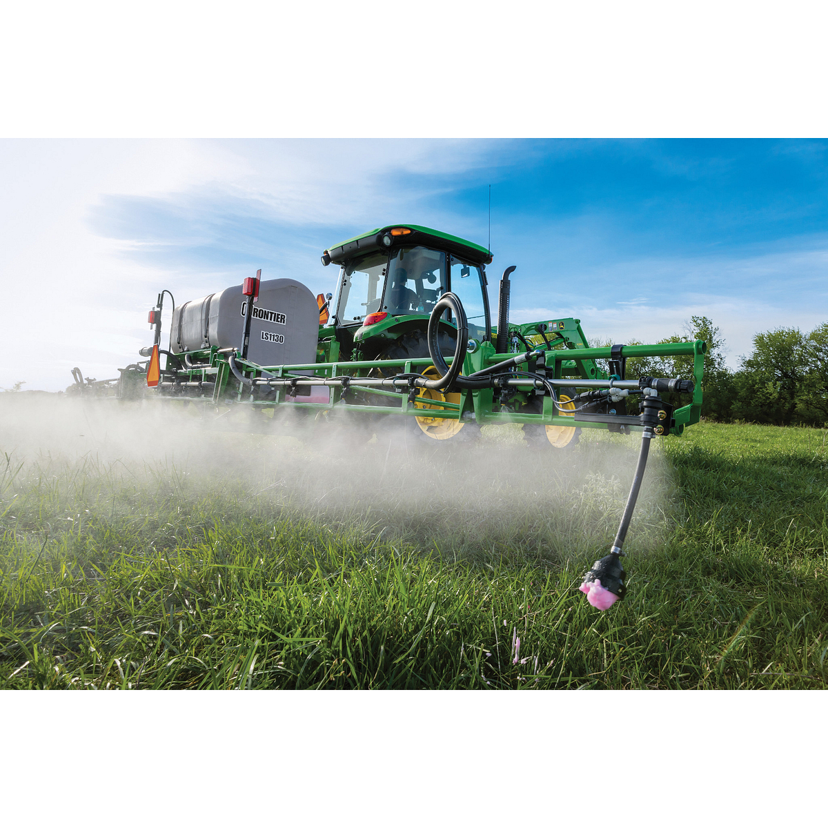 A agricultural sprayer tractor applies a fine mist over a grassy field using extended spray booms, with nozzles releasing chemicals close to the ground under a blue sky.