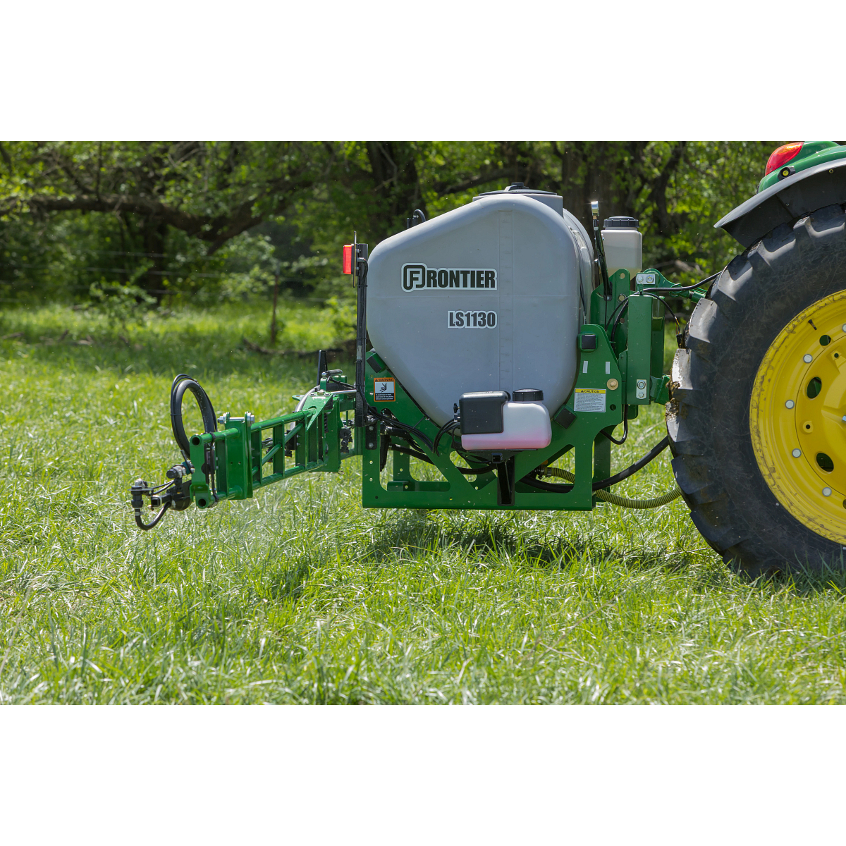 Frontier LS1130 tractor-mounted sprayer with a gray tank spraying attachment positioned on grass, connected to a tractor tire, outdoors in a grassy field with trees in the background