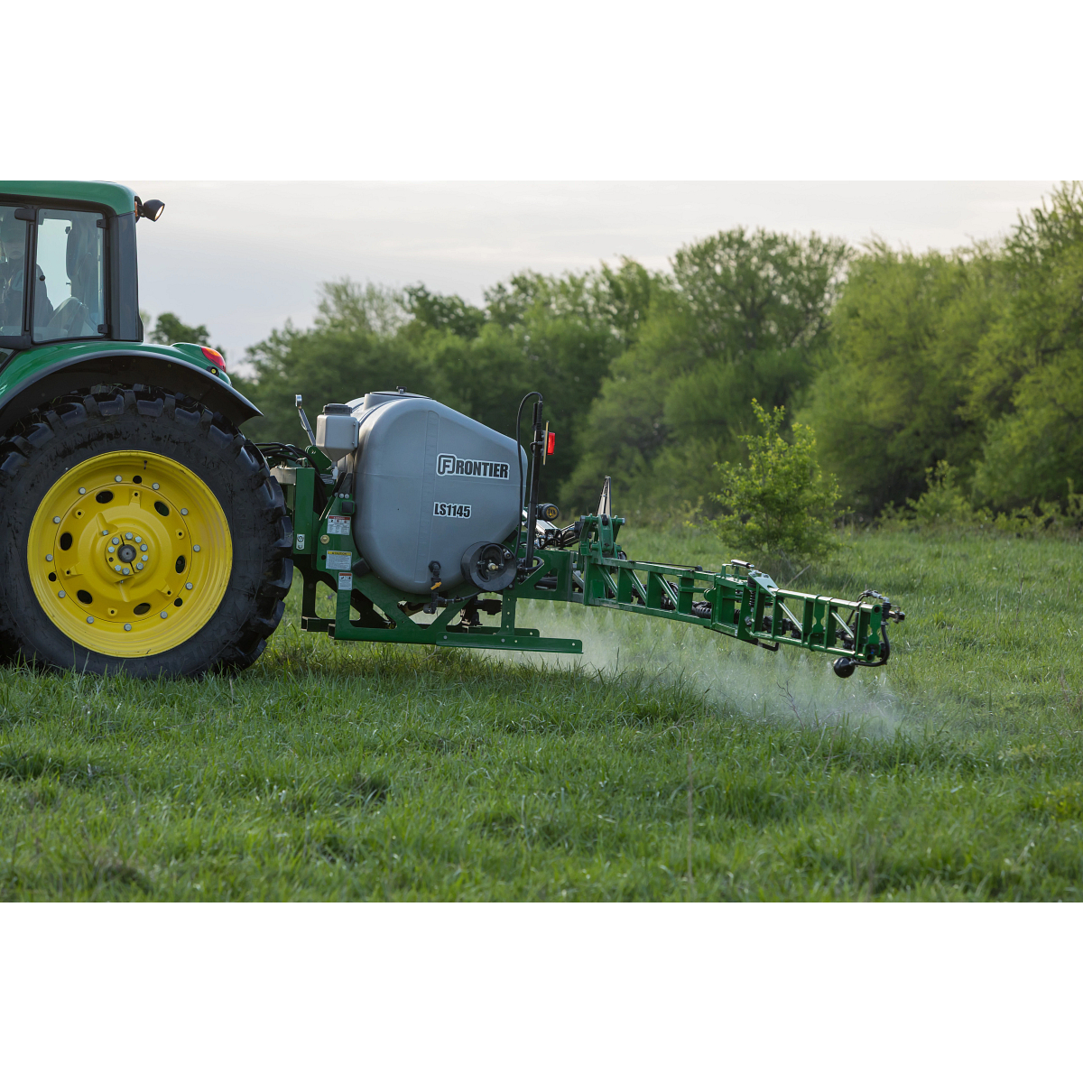 A tractor with yellow wheels pulls a mounted crop sprayer, spraying liquid over a grassy field, with trees in the background.