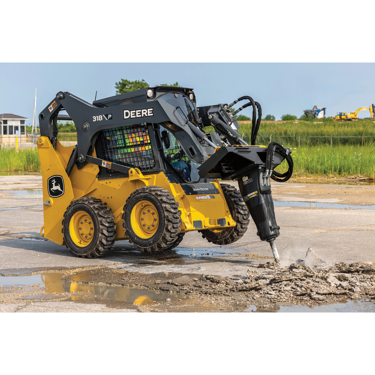 A 318 P-Tier skid steer operates on a paved surface, using a mounted hydraulic breaker attachment to break up concrete.
