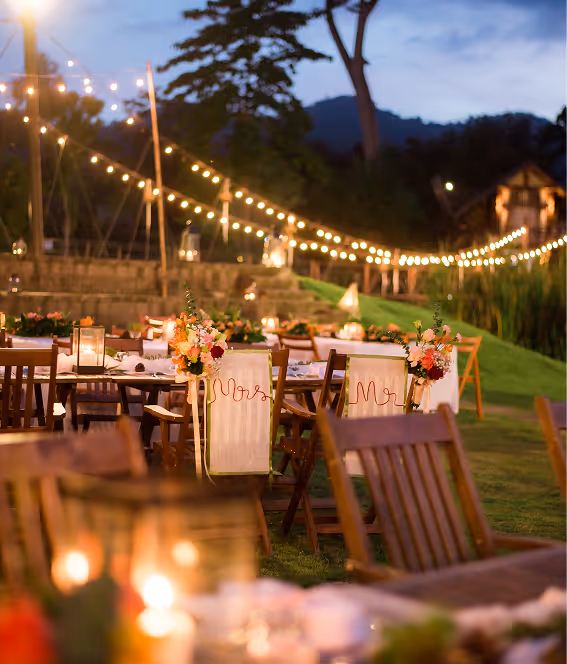Tables et chaises en bois décorées de fleurs avec des guirlandes lumineuses en arrière-plan lors d'une soirée en plein air.