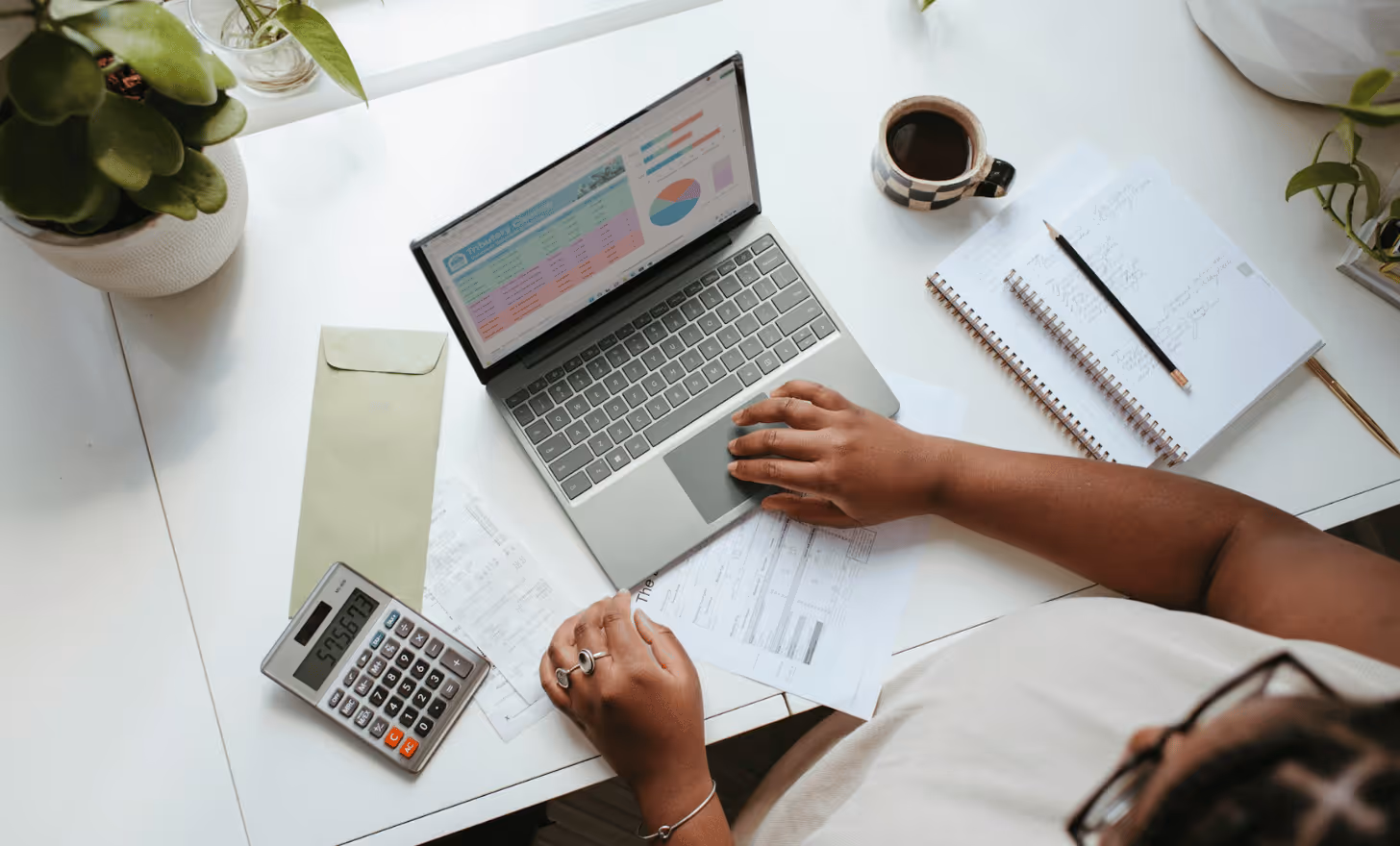 Personne utilisant un ordinateur portable avec un graphique et un tableau à l'écran, entourée de documents, calculatrice, cahiers, crayon, tasse de café et plantes sur un bureau blanc.