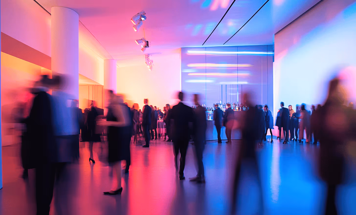 Personnes en tenue de soirée socialisant dans un espace intérieur éclairé par des lumières colorées rosées et bleues avec un bar en arrière-plan.