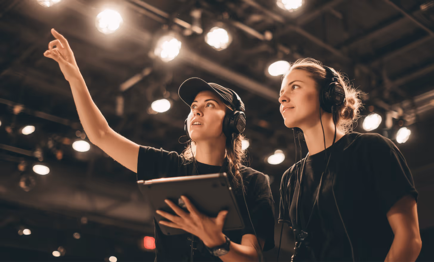 Deux femmes portant un casque audio discutent sous des projecteurs dans un studio, l’une tenant une tablette et pointant du doigt.
