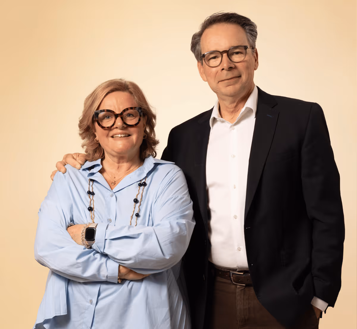 Un homme et une femme âgés souriants, l'homme porte un costume noir et la femme une chemise bleue avec des lunettes.