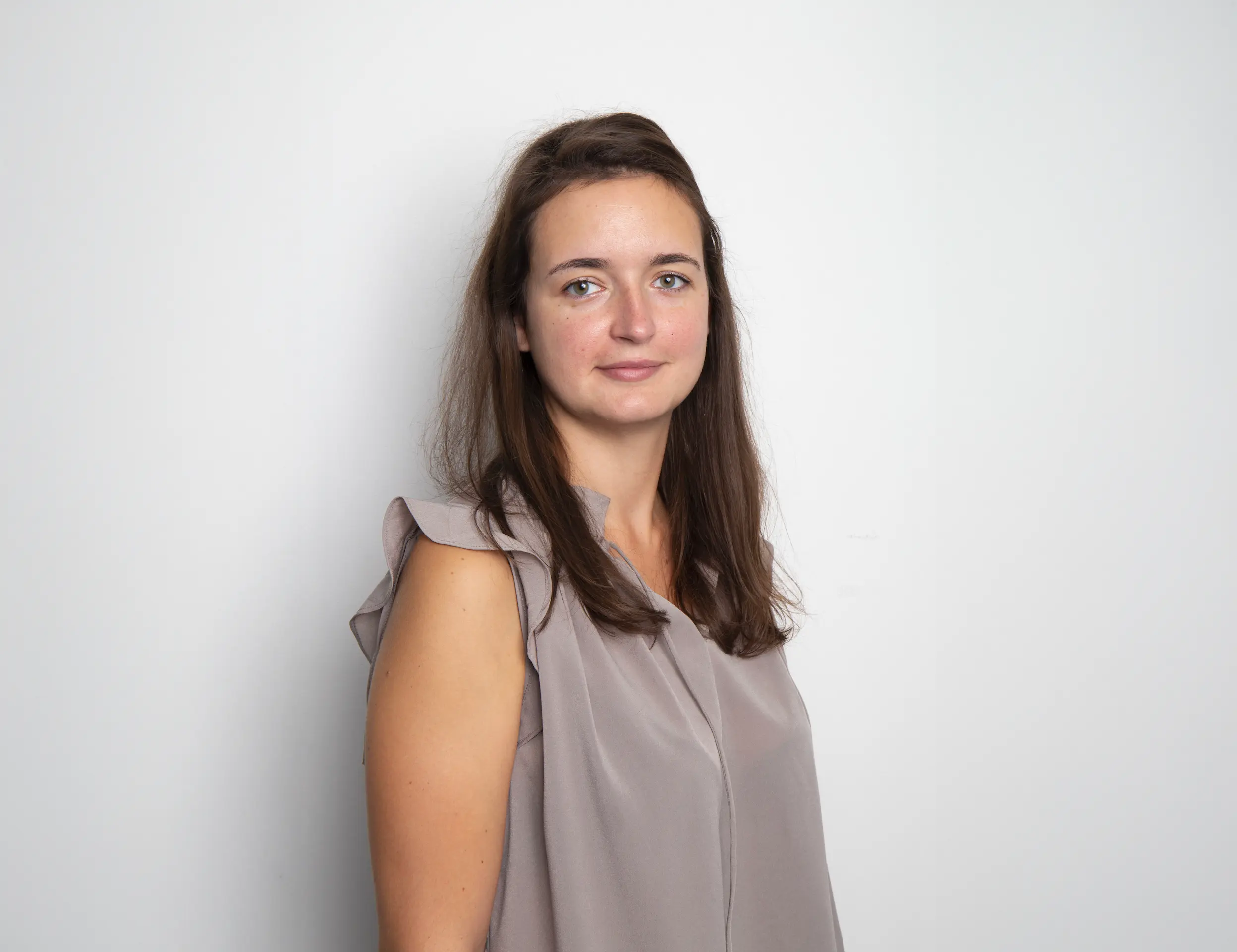 Young woman with long brown hair wearing a sleeveless beige blouse standing against a plain white background.