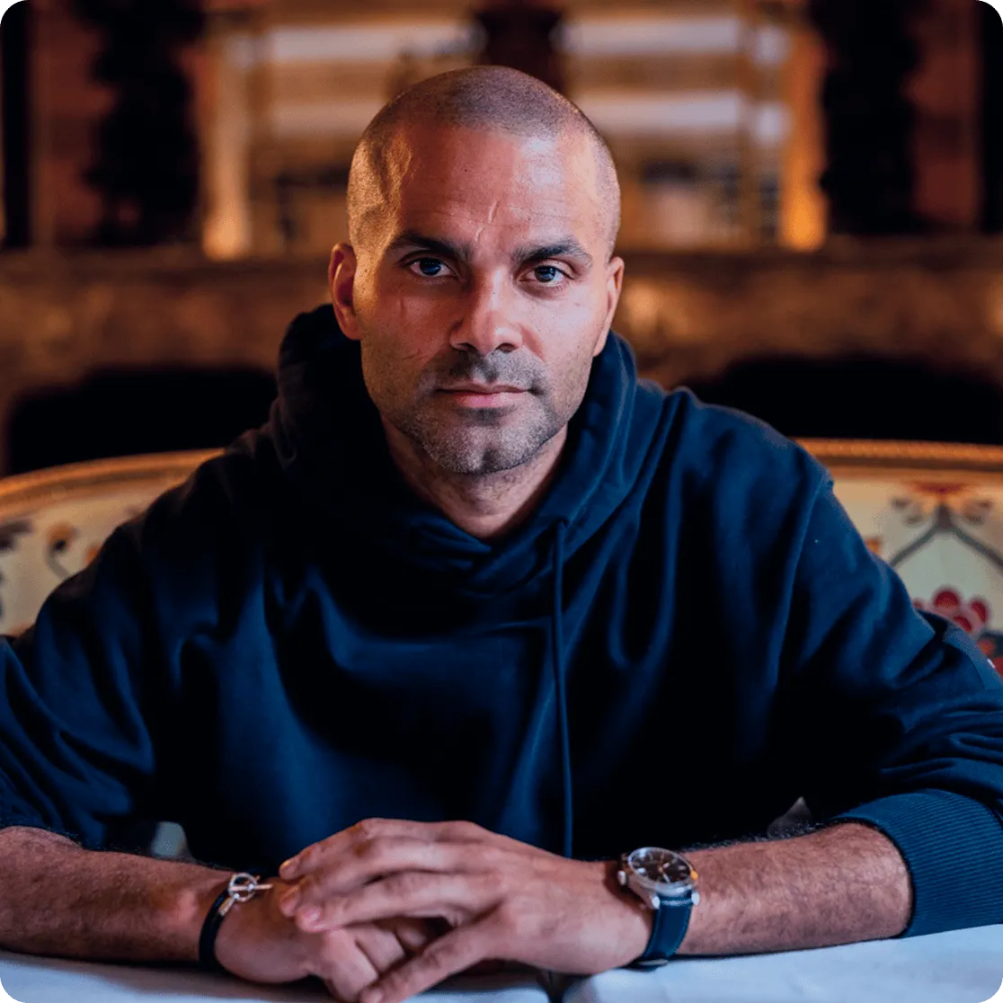 Tony Parker wearing a dark hoodie, sitting with hands clasped on a table in a warmly lit room.