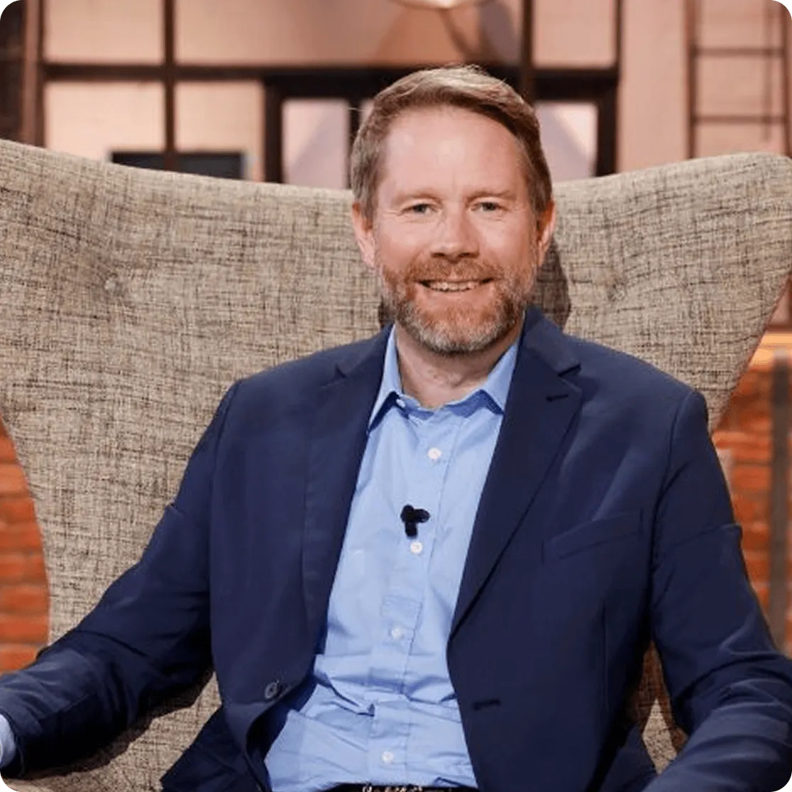 Éric Larchevêque wearing a blue suit jacket and light blue shirt sitting on a textured beige chair.