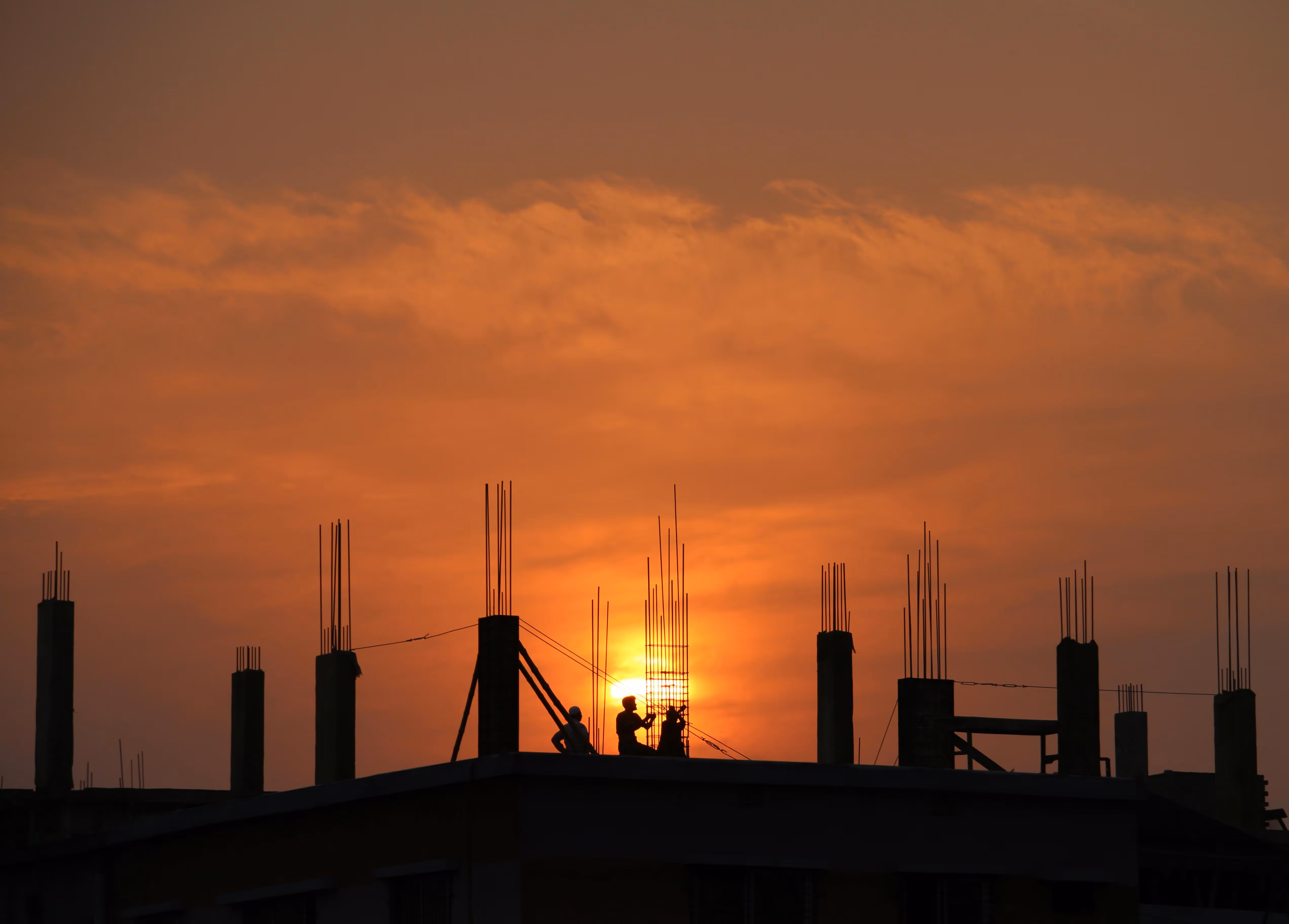 Lavoratori in silhouette su un cantiere al tramonto con il sole arancione sullo sfondo.