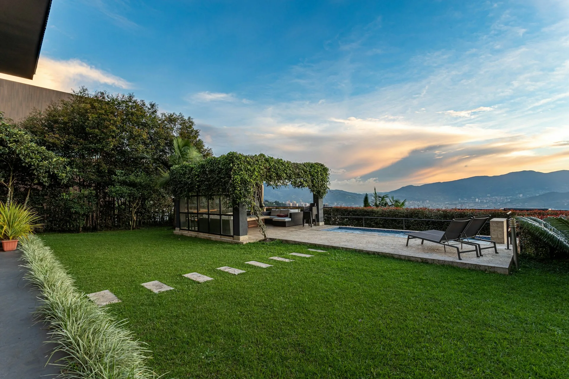 Backyard with green lawn, stone path, covered seating area with greenery, two lounge chairs by a pool, and mountains under a blue sky at sunset.