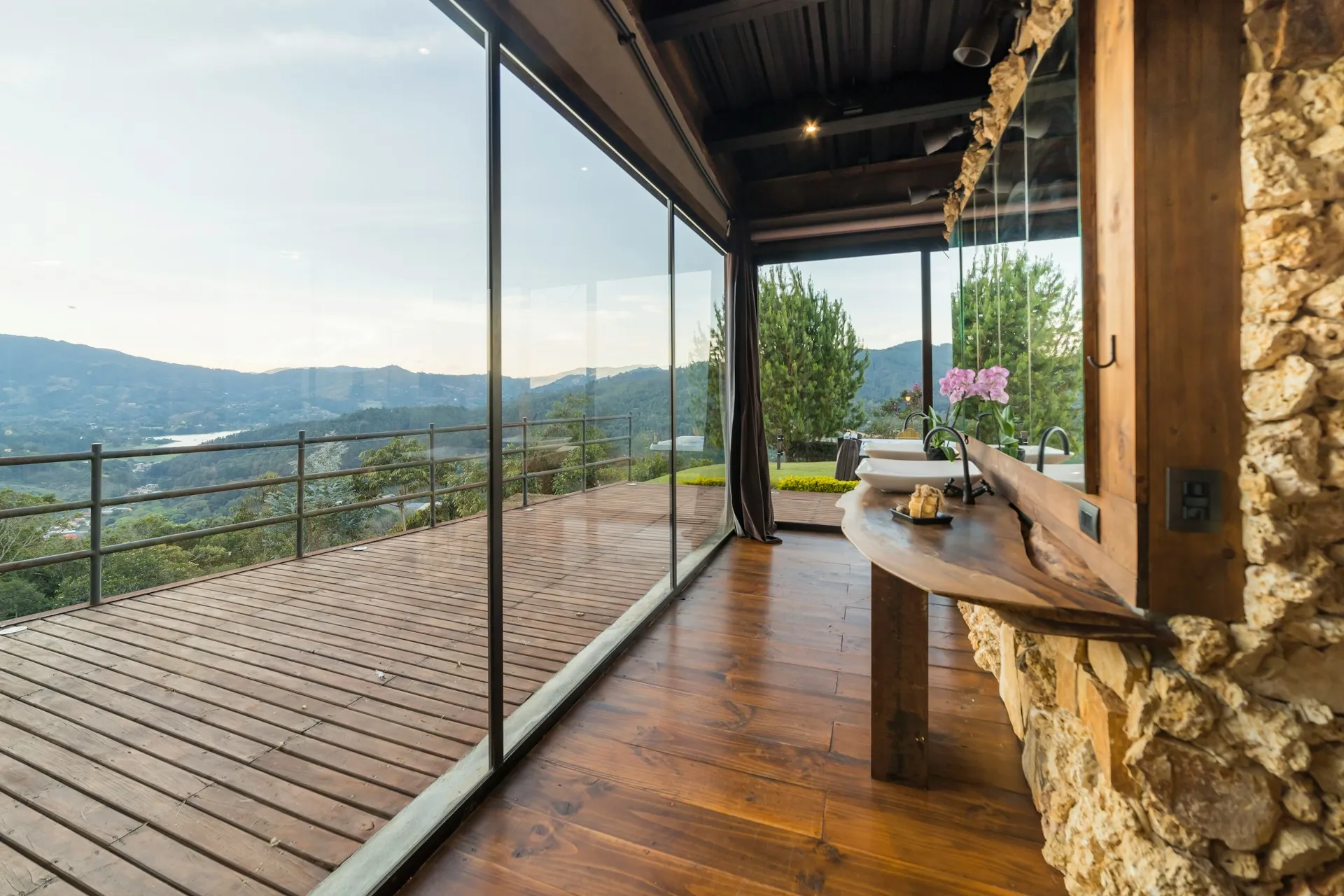 Interior view of a rustic bathroom with a wooden counter and vessel sinks overlooking a mountain landscape through floor-to-ceiling glass windows.