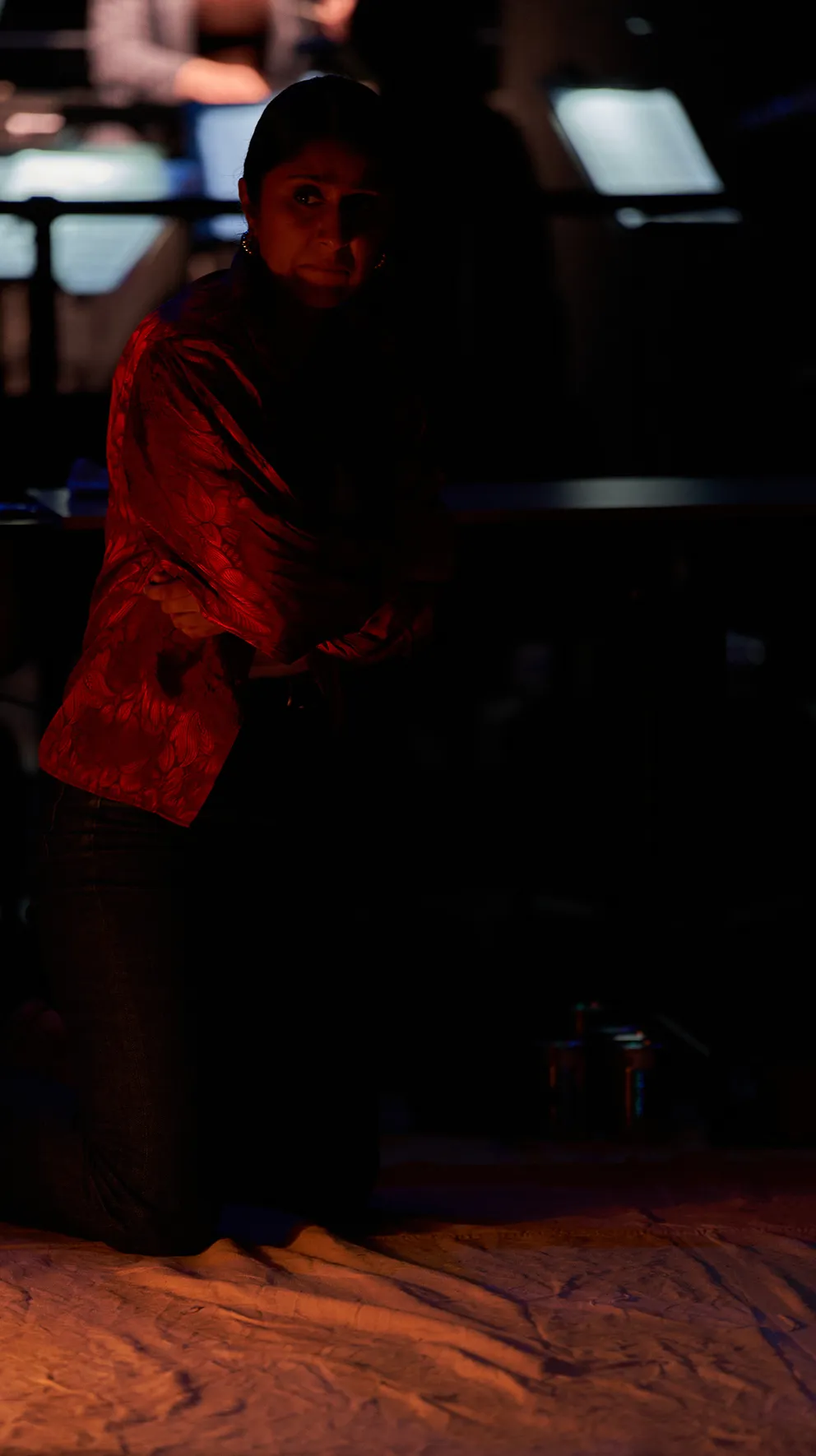 Woman kneeling on a dimly lit stage in a red patterned top.