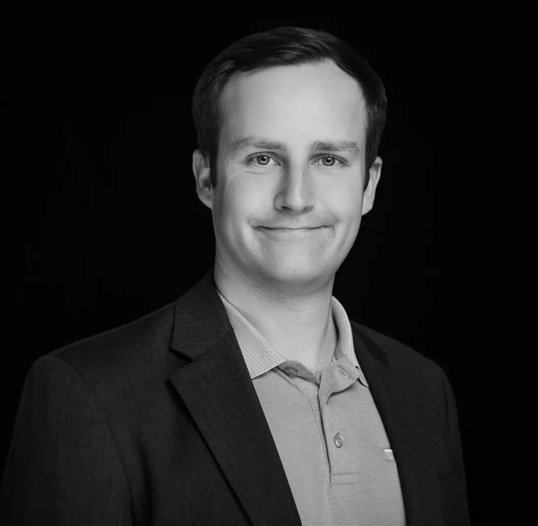 Portrait of a man in a suit jacket and polo shirt, smiling against a dark background.