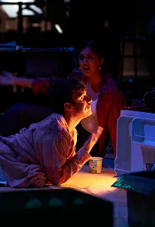 Actor lying on the floor under warm stage light while another performer gestures behind them.
