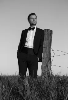 Man in a tuxedo leaning against a fence post in a grassy field.