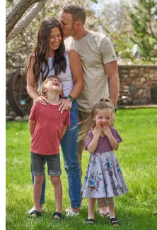 Family of four in a yard, with the father kissing the mother on the head while kids laugh.