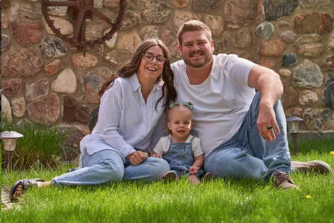 Parents sitting on the grass with their smiling toddler in front of a stone wall.