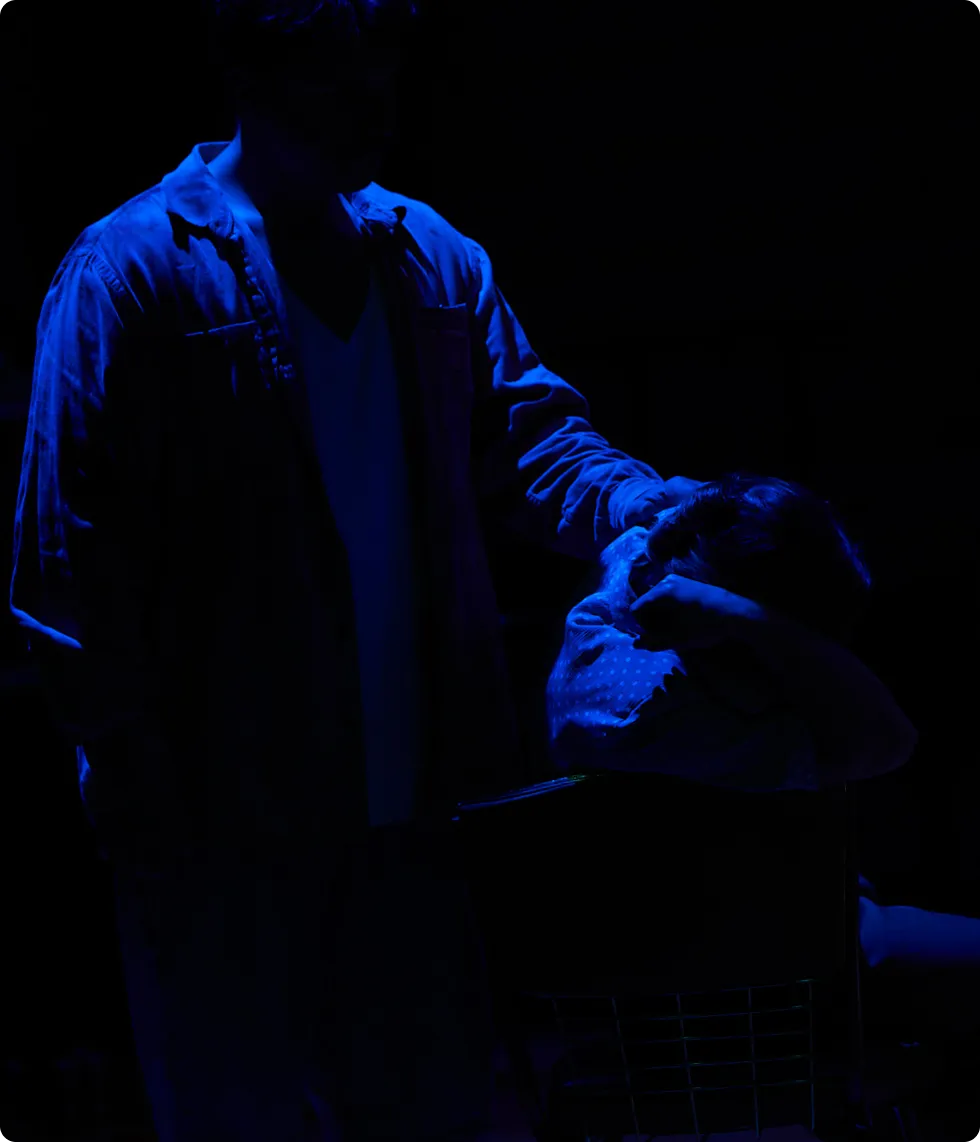 A man stands over a person lying on a low surface, dramatically lit from above by blue light in a dark scene.