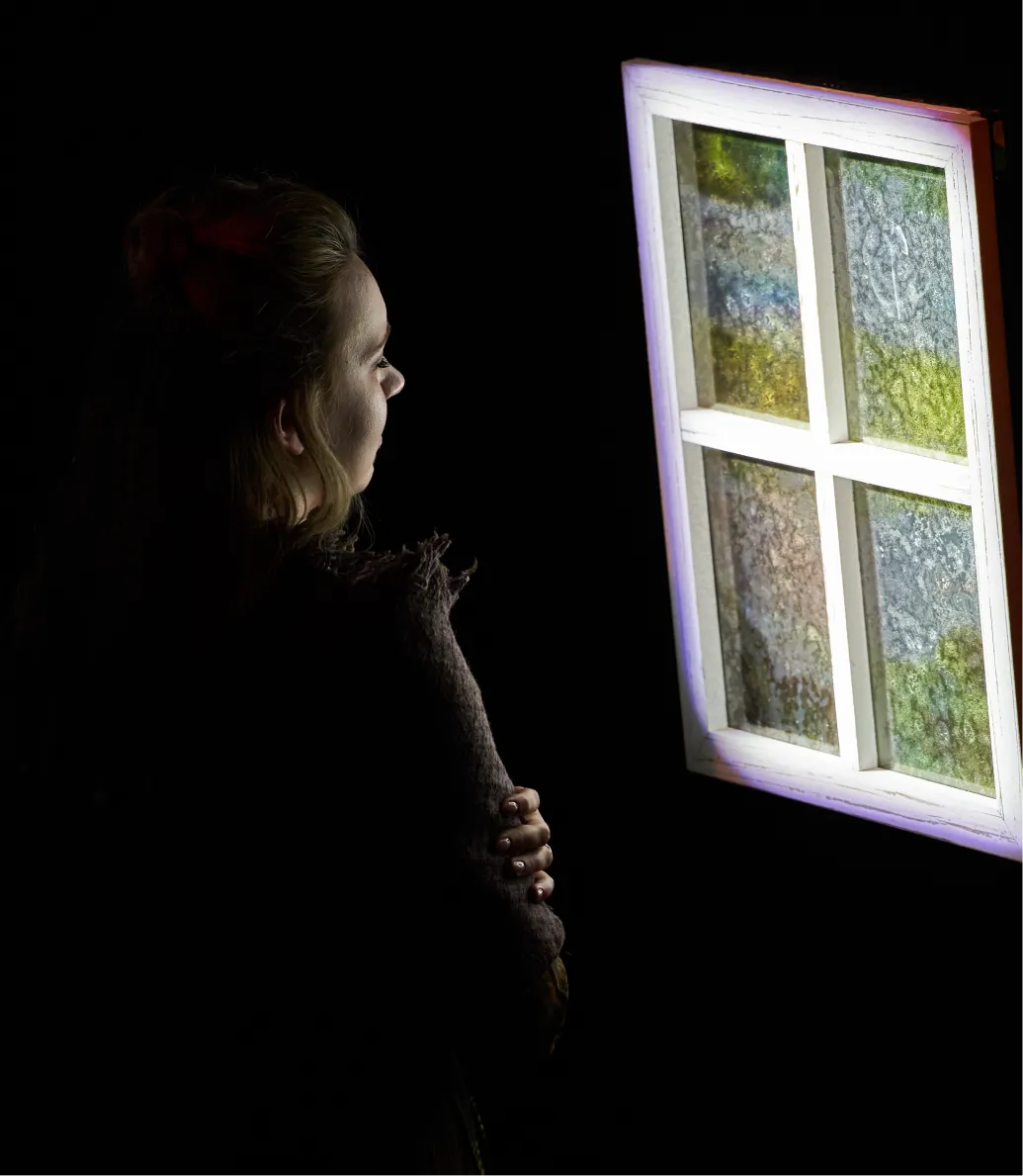 A woman in shadow looks toward a brightly lit window with frosted, colorful panes.