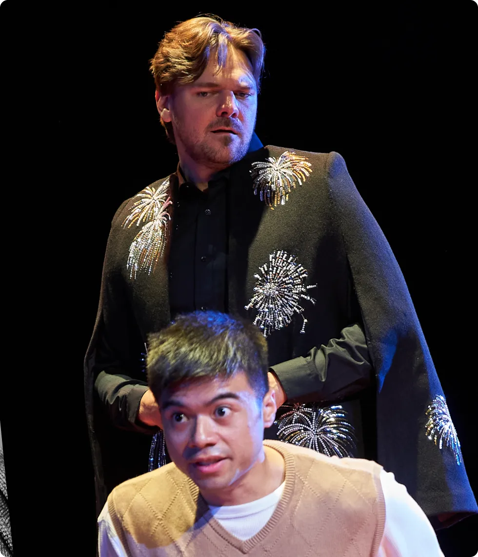 A man in a black cape embroidered with firework designs stands behind another man in a beige vest, both looking off-camera with intense expressions.