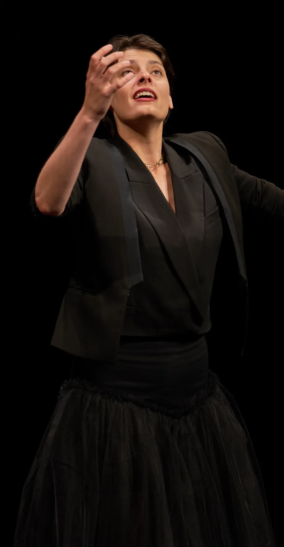 An actress wearing a black blazer and tutu skirt stands against a black background, looking up with her right hand raised dramatically.