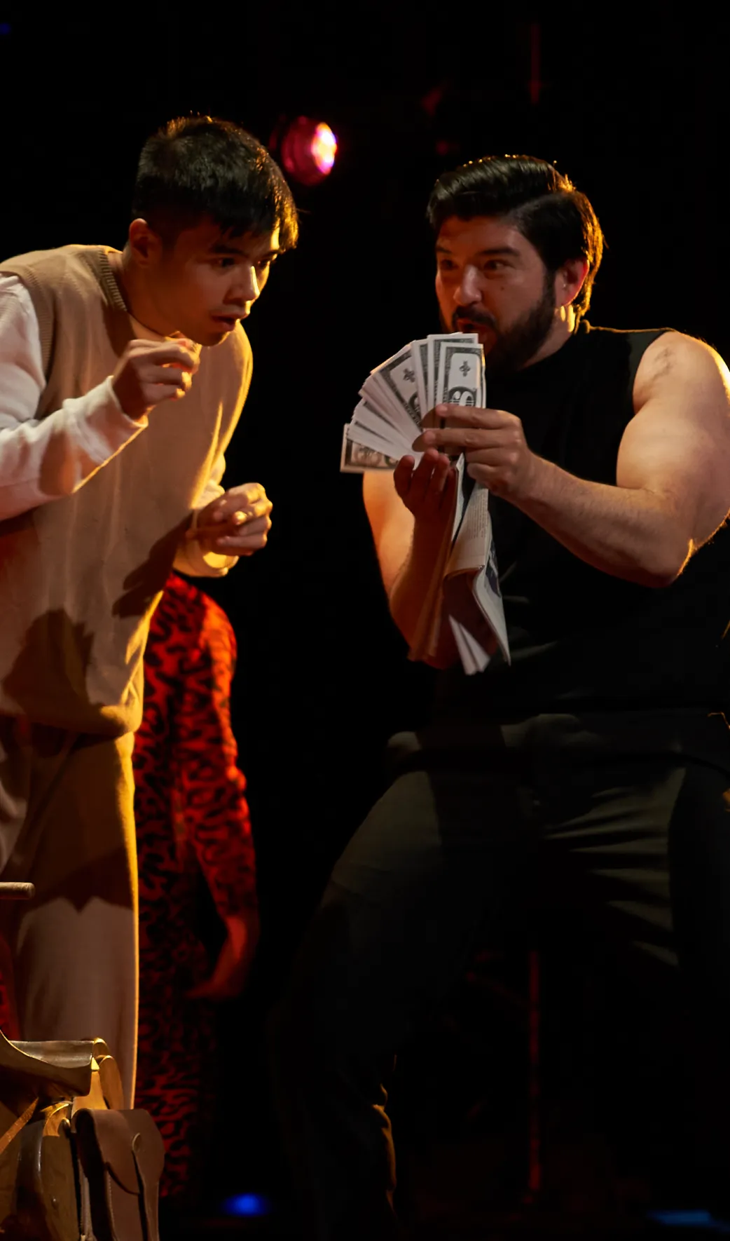 Two actors on a dark stage; one man in a black shirt holds up a fan of cash, while the other in beige clothing looks on intently.