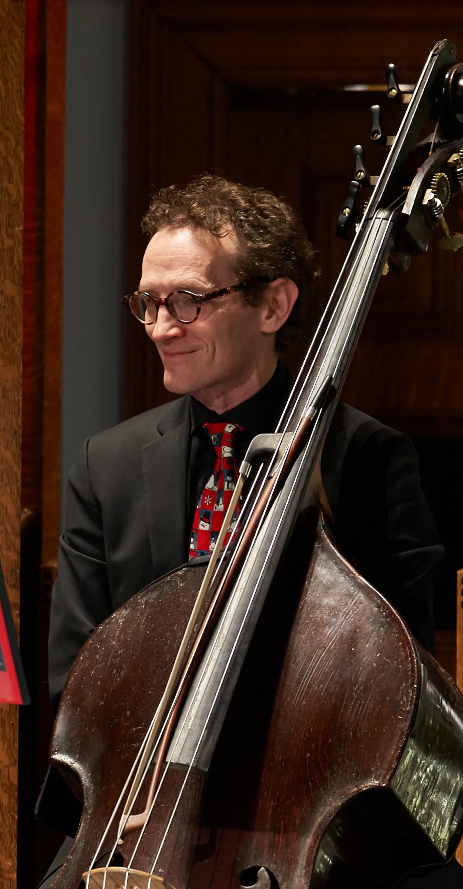 A man in glasses and a suit smiles while holding and standing behind an upright acoustic bass.