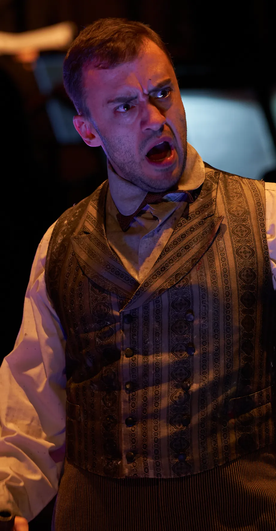 A man in a period vest and shirt on stage, illuminated in dark lighting, shouts with an intense, angry expression.