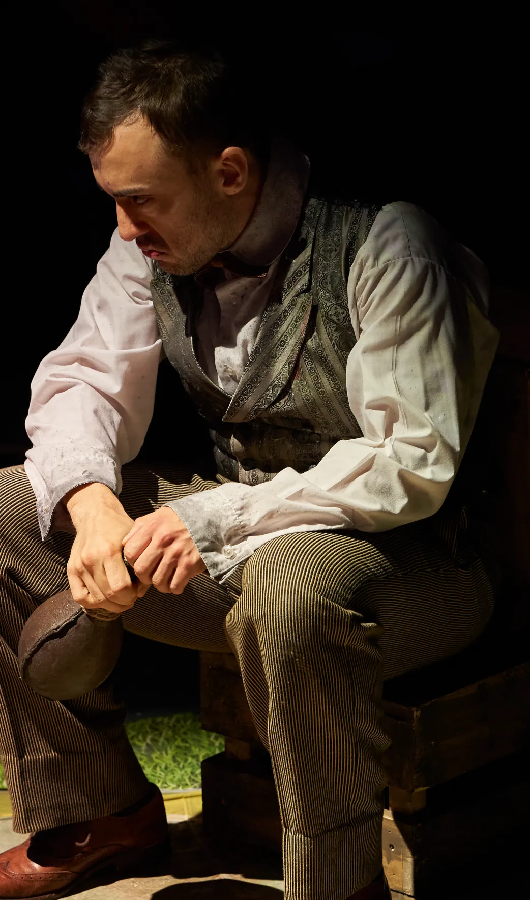 A man in historical clothing sits in the dark, holding a dark object with a focused, serious expression.