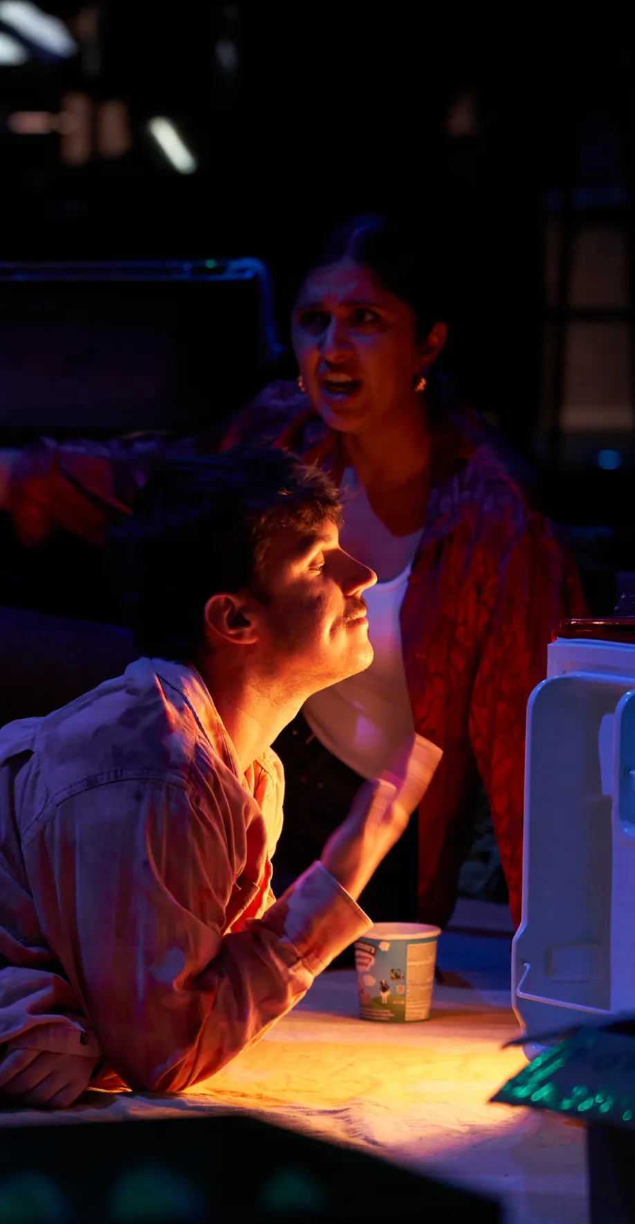 Two performers in a dramatic, dark stage light; a man looks up, and a woman appears agitated behind him.