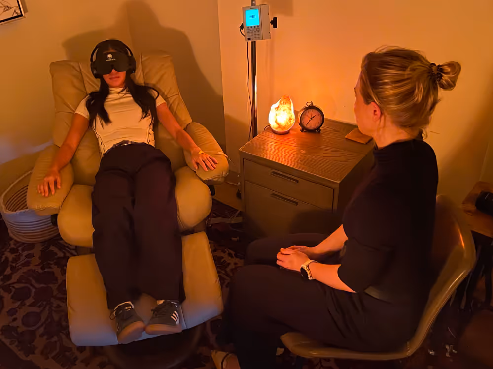 Woman relaxing in a recliner with headphones and an eye mask during a therapy session, while another woman sits nearby in a warmly lit room with a salt lamp and clock on a table.