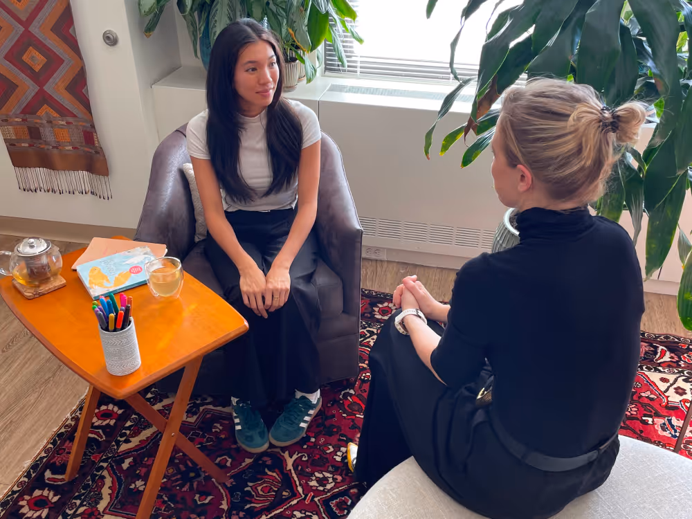Two women sitting across from each other in a cozy office setting, engaged in conversation, with plants and natural light from a window in the background.