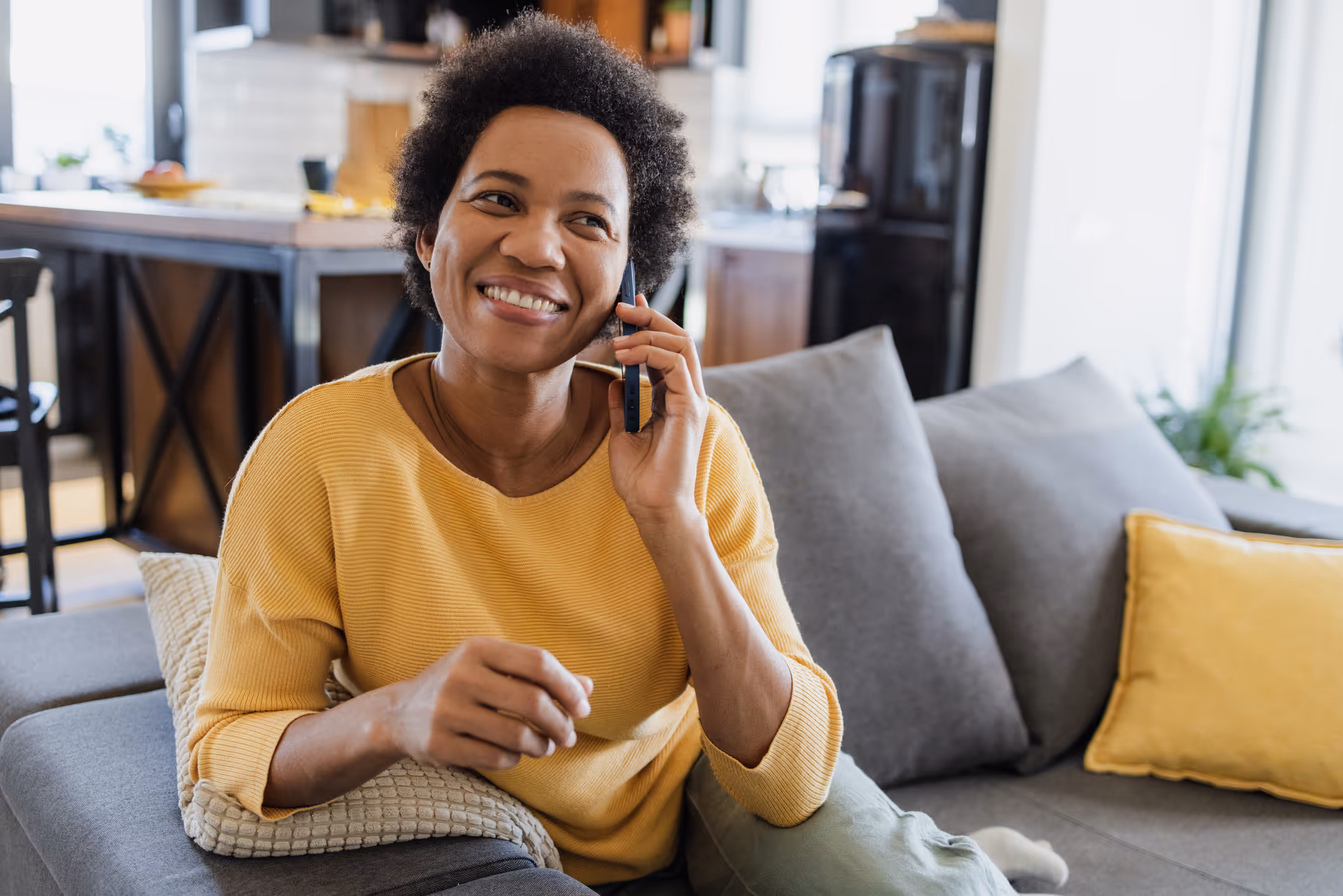 Smiling woman sitting on a couch at home, wearing a yellow sweater and talking on the phone, with a cozy living room and kitchen in the background.