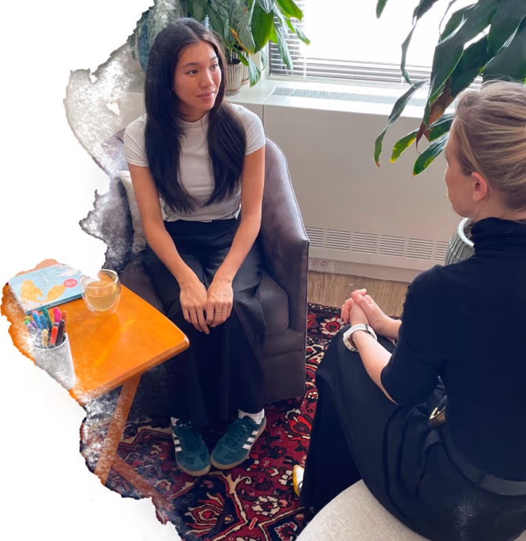 Two women sitting across from each other in a cozy office setting, engaged in conversation, with plants and natural light from a window in the background.