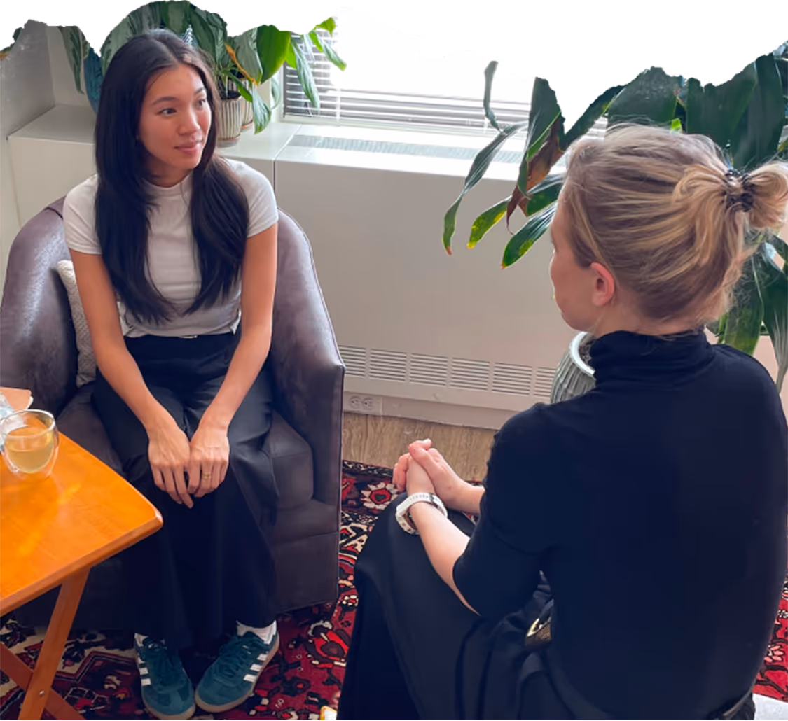 Two women sitting across from each other in a cozy office setting, engaged in conversation, with plants and natural light from a window in the background.