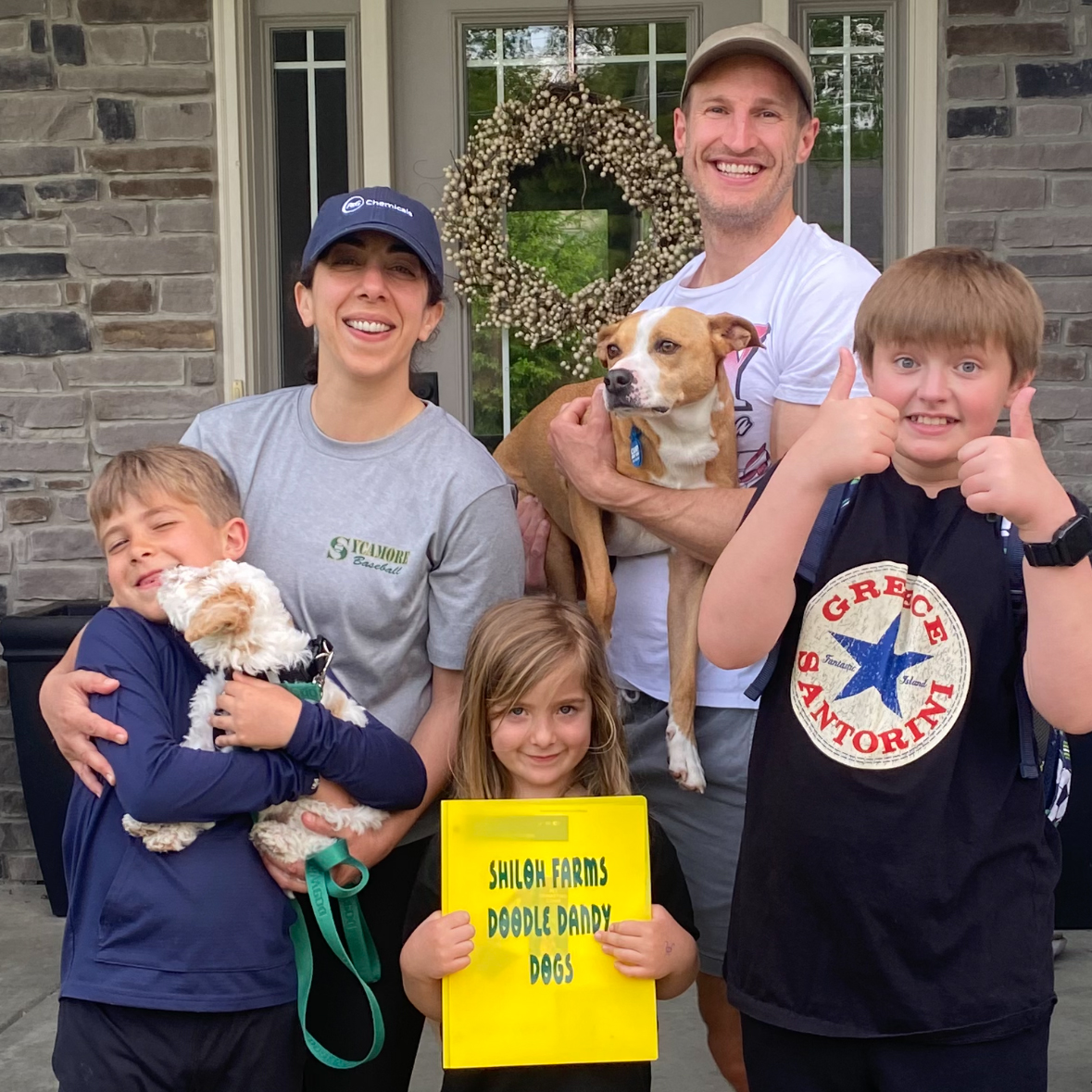Smiling family of two adults and three children holding two dogs and a yellow sign reading 'Shiloh Farms Doodle Dandy Dogs' in front of a house door with a wreath.