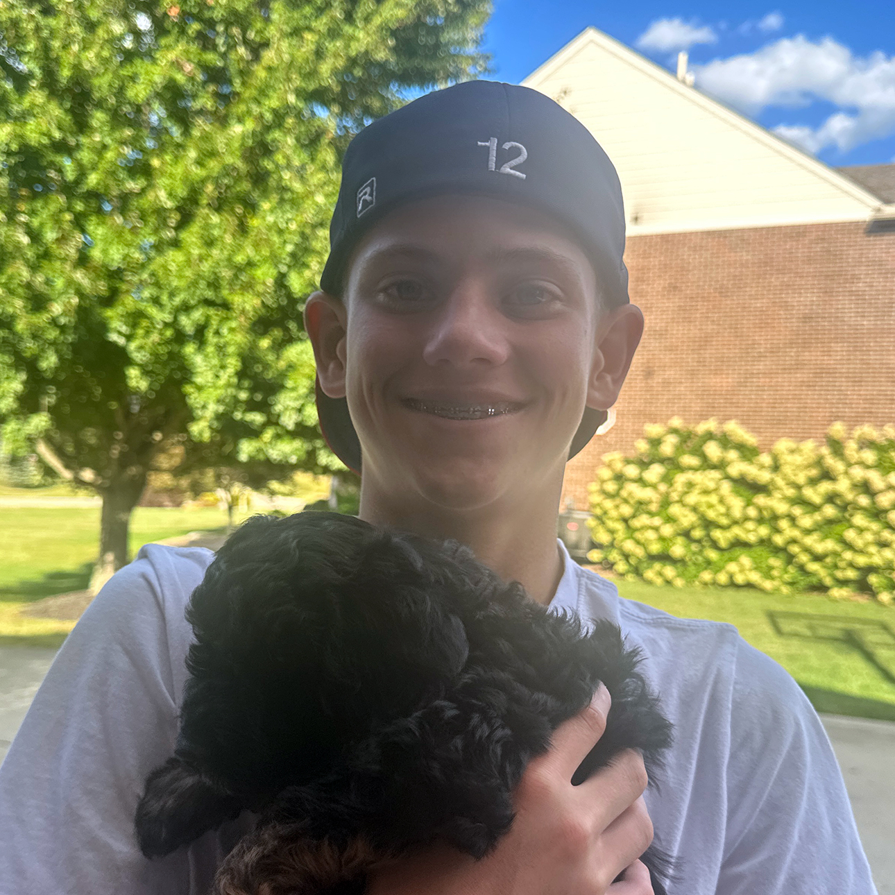Smiling boy wearing a backward black cap holding a small black dog outdoors with trees and a house in the background.