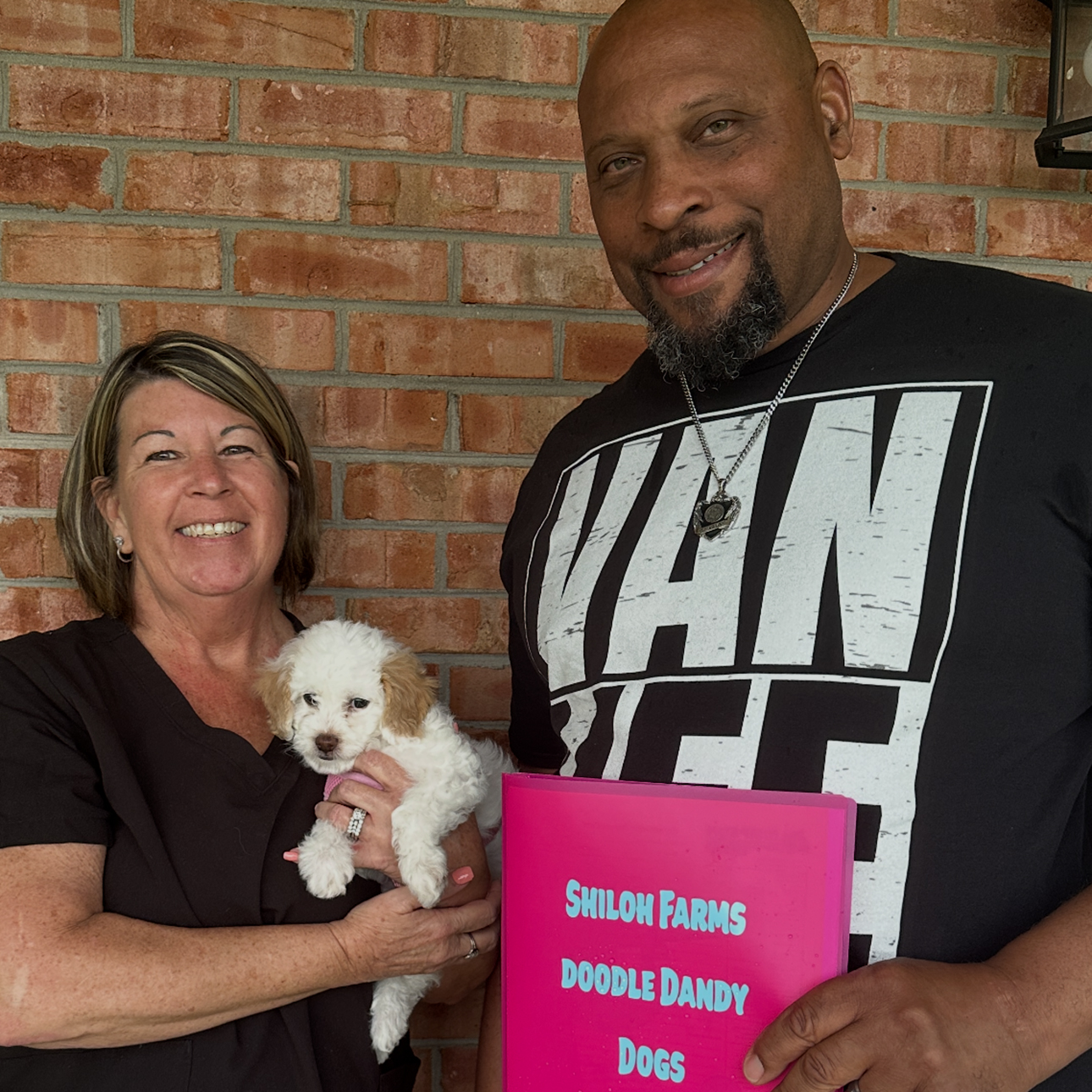 Smiling woman holding a small white and tan puppy next to a man holding a pink sign that reads 'Shiloh Farms Doodle Dandy Dogs' against a brick wall background.