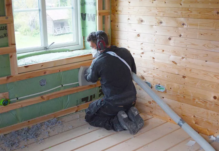 Medarbetare på Lösullsteamet som  sprutar lösullsisolering i väggen på ett trähus med skyddsmask och hörselskydd.