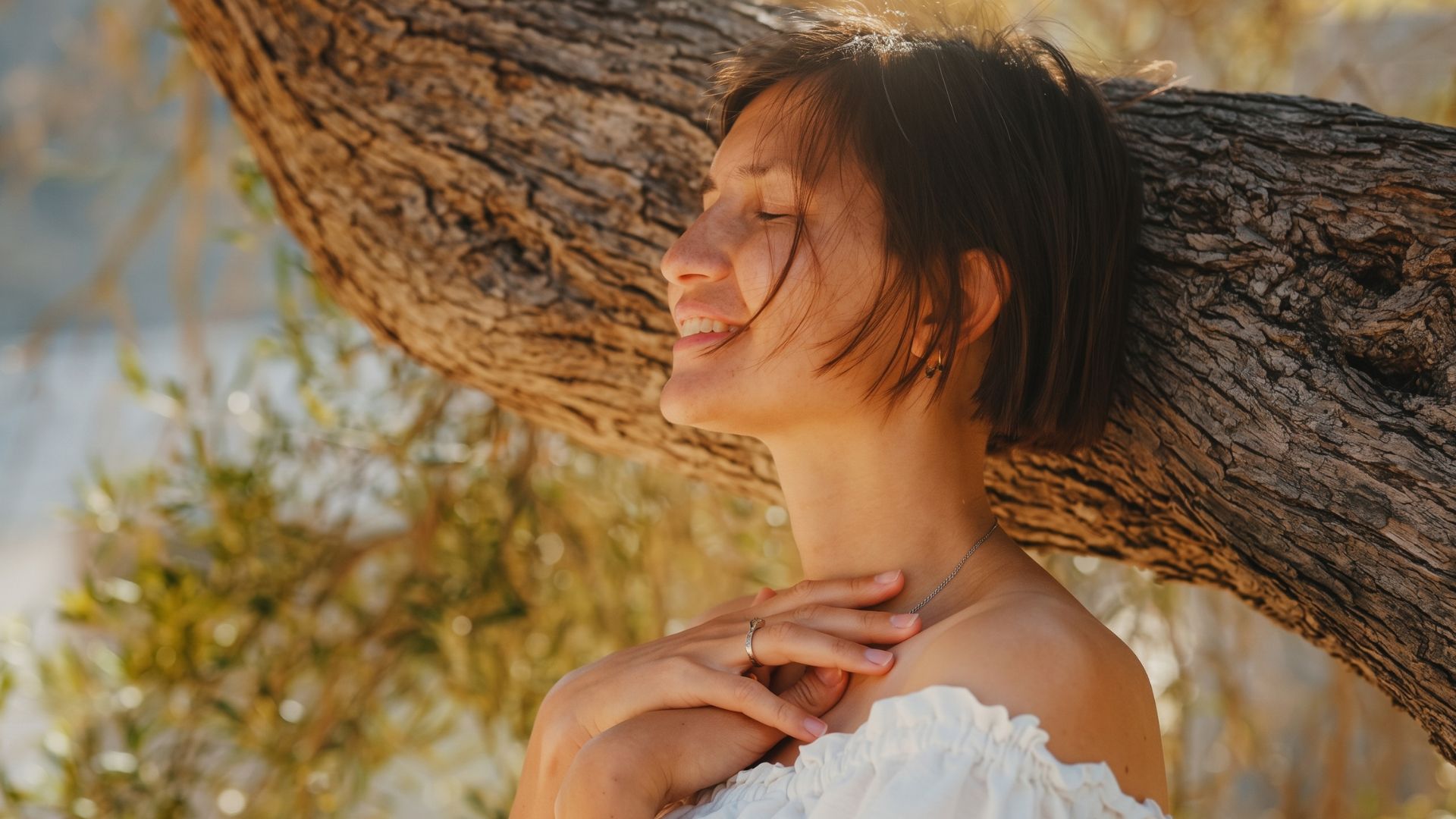 Woman with short dark hair leaning against a tree trunk outdoors, eyes closed and smiling gently, wearing a white off-shoulder top with hands touching her chest.
