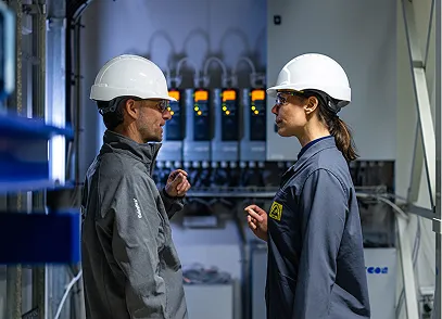 Two engineers wearing white hard hats and safety glasses having a discussion in an industrial control room.