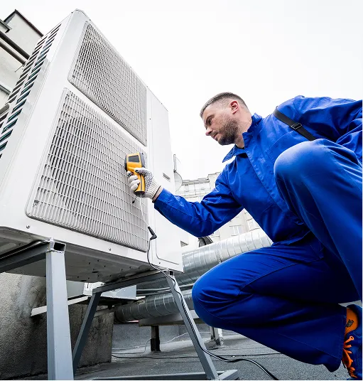 Technician in blue coveralls using a handheld device to check an outdoor air conditioning unit on a rooftop.