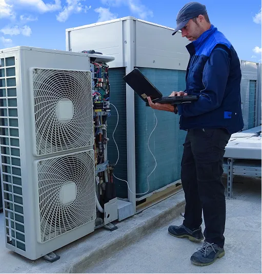 Technician in blue uniform using a laptop while inspecting an outdoor air conditioning unit on a rooftop under a clear sky.