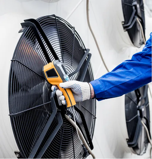 Gloved hand in blue work uniform holding a yellow infrared thermometer pointed at a large black industrial fan.