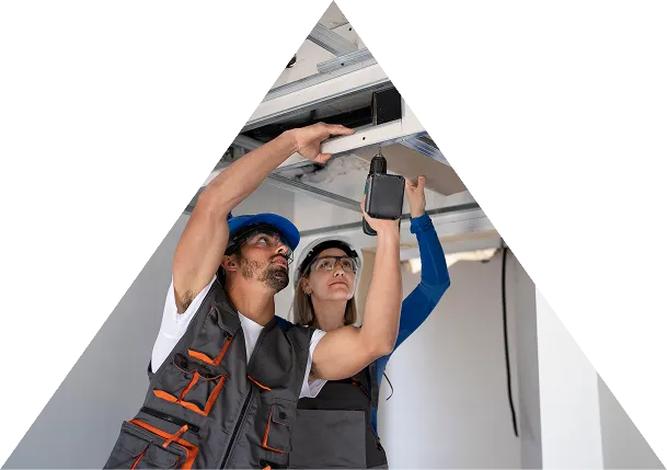 Two construction workers wearing safety gear installing a metal frame on a ceiling using a power drill.