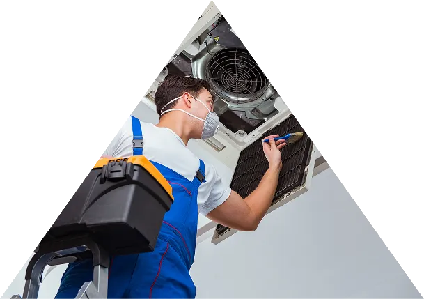 Technician wearing a face mask and blue overalls repairing an air conditioning vent inside a building ceiling.