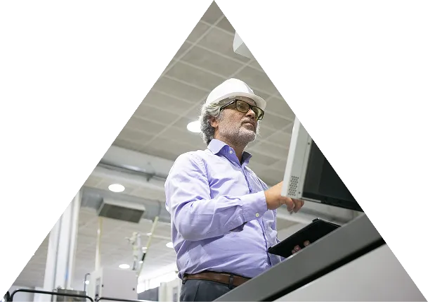 Man in a white hard hat and glasses working with a computer terminal in an industrial or manufacturing setting.