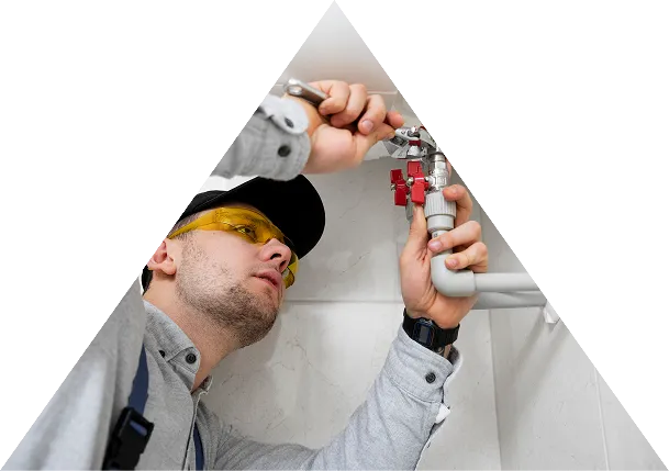 Plumber wearing safety glasses and a cap fixing pipes under a sink using a wrench.