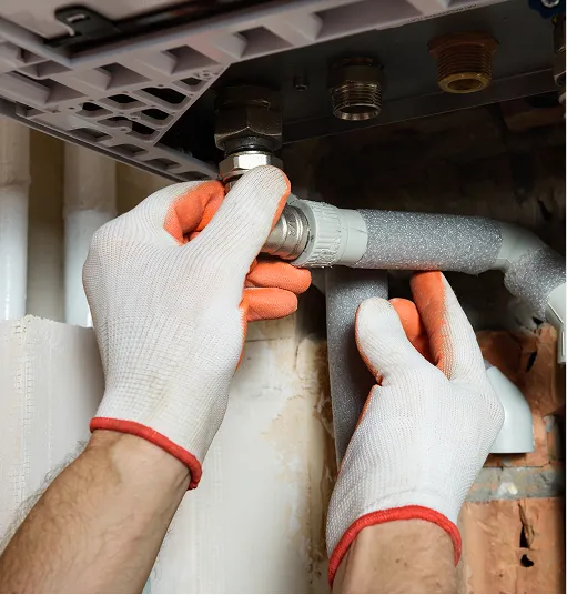 Hands wearing white gloves with orange grip installing or tightening a plumbing pipe connection under a sink.